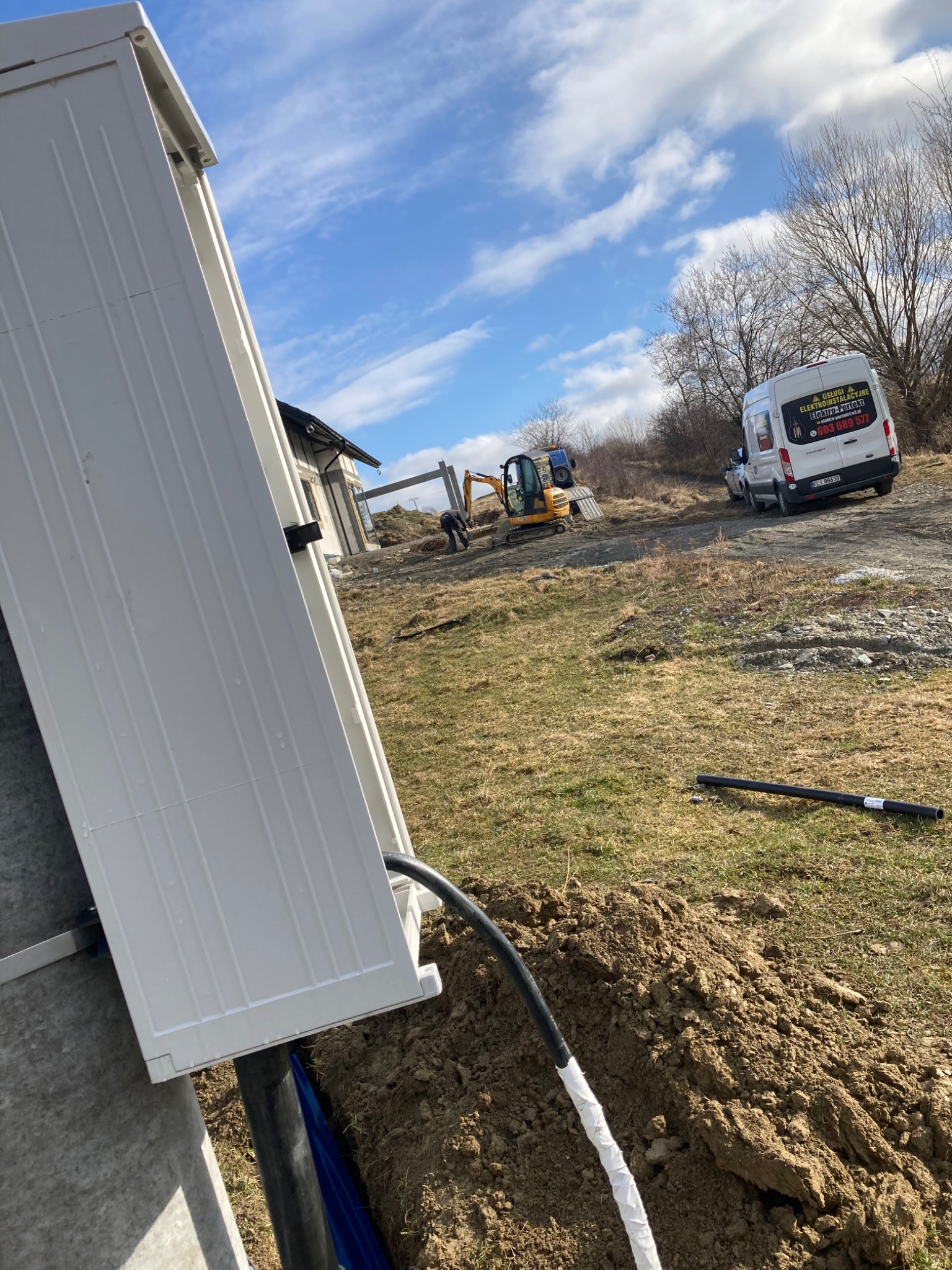 Skrzynka elektryczna z wyprowadzonymi kablami w ziemi, w tle koparka i samochód z napisem 'Usługi Elektroinstalacyjne', błękitne niebo z chmurami.