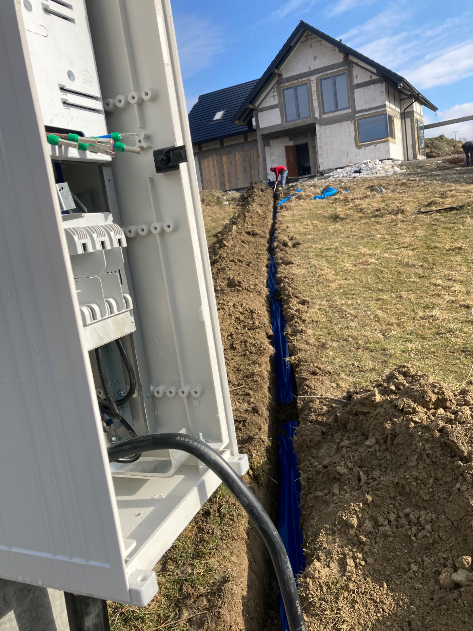 Skrzynka elektryczna z otwartymi drzwiczkami, widoczne kable i złącza, w tle wykopany rów z niebieską rurą osłonową oraz budynek w stanie surowym otwartym.