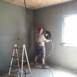 Pracownik w szortach wygładza świeżo nałożony tynk na ścianie za pomocą elektrycznej szlifierki, widoczna drewniana drabina i surowy sufit z płyty OSB.