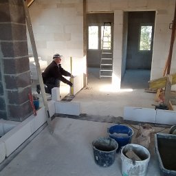 WeSo Projekt - Wnętrze poddasza w trakcie budowy ścian działowych z bloczków, widoczny murarz z poziomicą, surowe ściany, drewniana konstrukcja dachu, narzędzia budowlane i worki z zaprawą.