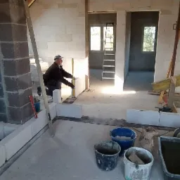 Wnętrze poddasza w trakcie budowy ścian działowych z bloczków, widoczny murarz z poziomicą, surowe ściany, drewniana konstrukcja dachu, narzędzia budowlane i worki z zaprawą.