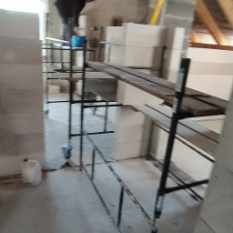 WeSo Projekt - Wnętrze w trakcie budowy, widoczne rusztowanie, bloczki Ytong i murarz w tle, budujący ściany działowe na poddaszu z drewnianą konstrukcją dachu.