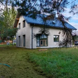 Surowy, dwukondygnacyjny dom z ciemną dachówką, jeszcze bez elewacji, otoczony świeżo skoszoną trawą i drzewami.