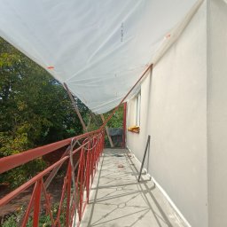 WeSo Projekt Grzegorz Miklasiewicz - Balkon zabezpieczony białą folią malarską, widoczne czerwone barierki, narzędzia malarskie i świeżo pomalowana ściana budynku w neutralnym odcieniu.