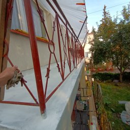 WeSo Projekt Grzegorz Miklasiewicz - Malowanie balustrady balkonowej na zewnątrz budynku, widoczna ręka z szmatką czyszcząca świeżą warstwę farby, przygotowane rusztowanie i zabezpieczenie folią malarską.