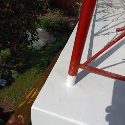 WeSo Projekt Grzegorz Miklasiewicz - Świeżo pomalowana na biało balustrada balkonu z czerwonymi metalowymi elementami, widoczne ślady farby na dolnych fragmentach nóg balustrady, w tle ogród z drzewami owocowymi i folią.