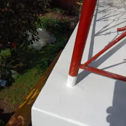 Świeżo pomalowana na biało balustrada balkonu z czerwonymi metalowymi elementami, widoczne ślady farby na dolnych fragmentach nóg balustrady, w tle ogród z drzewami owocowymi i folią.