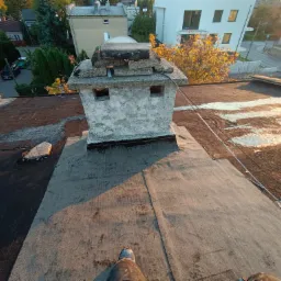 Widok z góry na płaski dach pokryty papą, z betonowym kominem z uszkodzeniami, częściowo naprawiony, w tle zabudowa mieszkalna i drzewa w jesiennym słońcu.