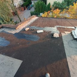 WeSo Projekt Grzegorz Miklasiewicz - Uszczelnianie dachu smołą wokół komina, widoczne cegły i fragment anteny satelitarnej na tle zabudowy miejskiej i zieleni.