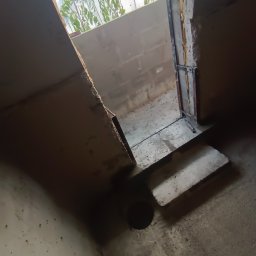 WeSo Projekt Grzegorz Miklasiewicz - Surowa piwniczka w trakcie hydroizolacji: widoczny otwór w posadzce, betonowe schody wejściowe oraz rośliny w donicach widoczne przez otwór w suficie.