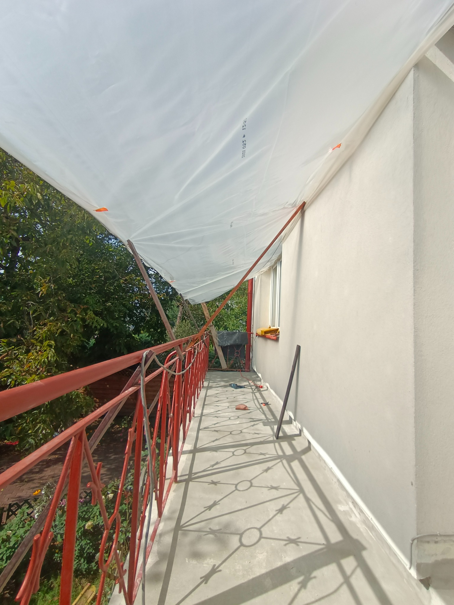 Balkon zabezpieczony białą folią malarską, widoczne czerwone barierki, narzędzia malarskie i świeżo pomalowana ściana budynku w neutralnym odcieniu.