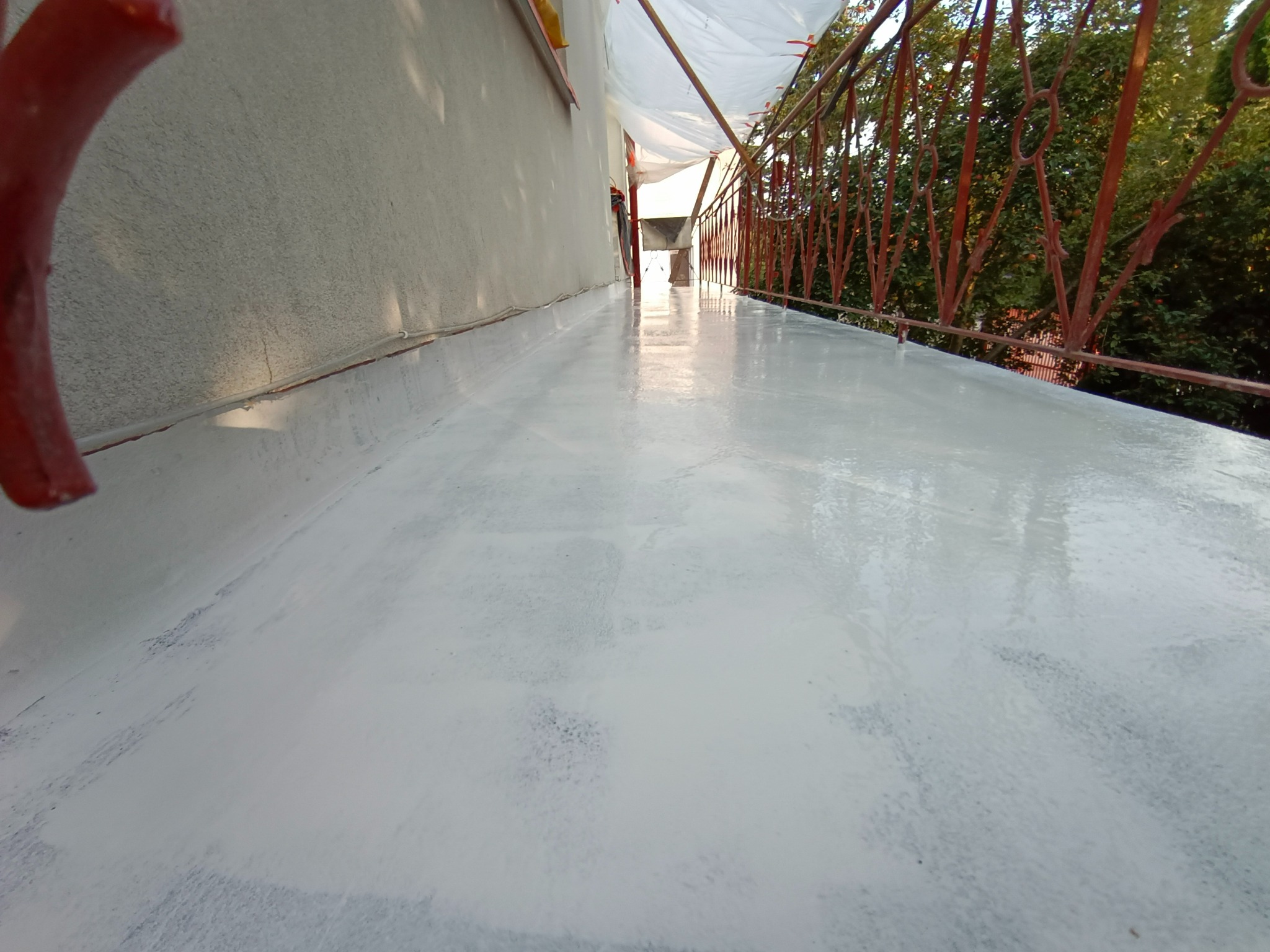 Świeżo pomalowana na biało podłoga balkonu z widoczną czerwoną barierką i zielenią drzew w tle.