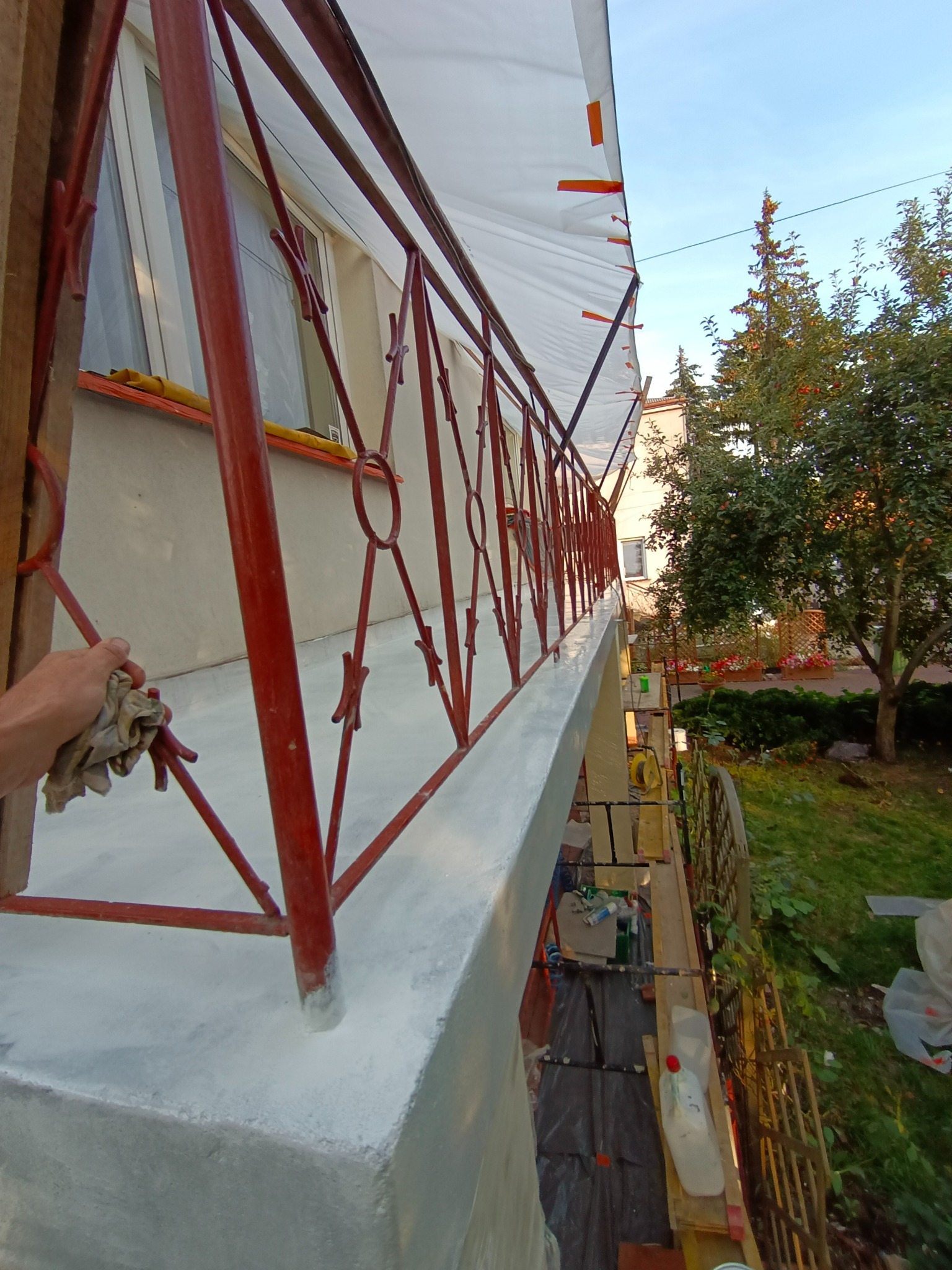 Malowanie balustrady balkonowej na zewnątrz budynku, widoczna ręka z szmatką czyszcząca świeżą warstwę farby, przygotowane rusztowanie i zabezpieczenie folią malarską.