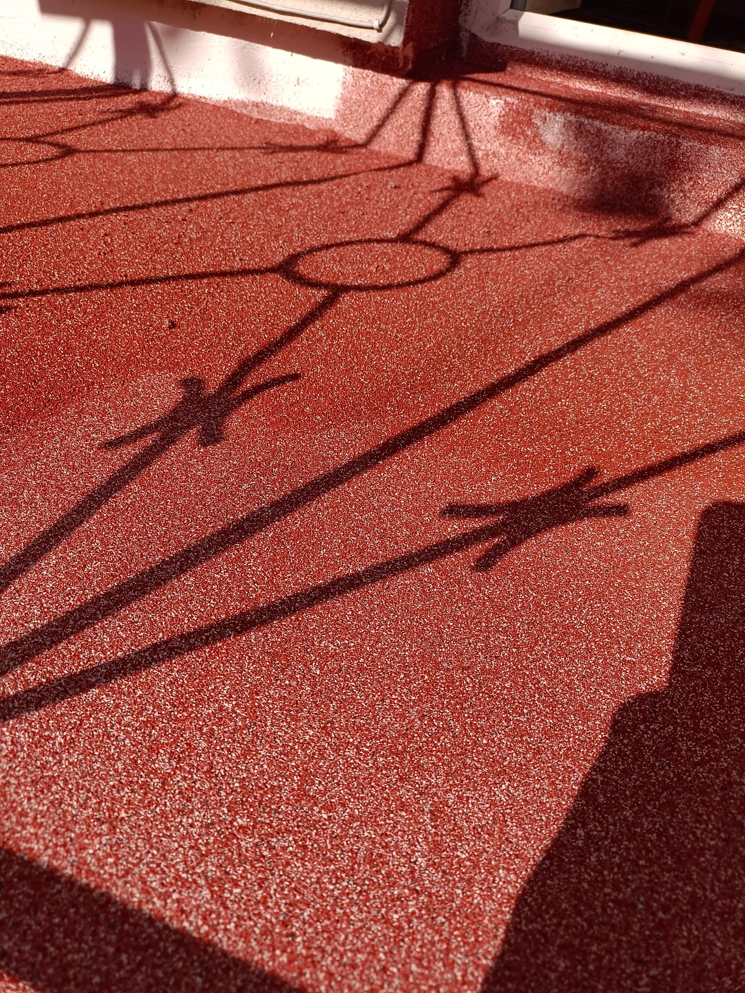 Czerwona, teksturowana powierzchnia dachu z widocznymi cieniami metalowej konstrukcji okiennej, oświetlona jasnym światłem słonecznym.