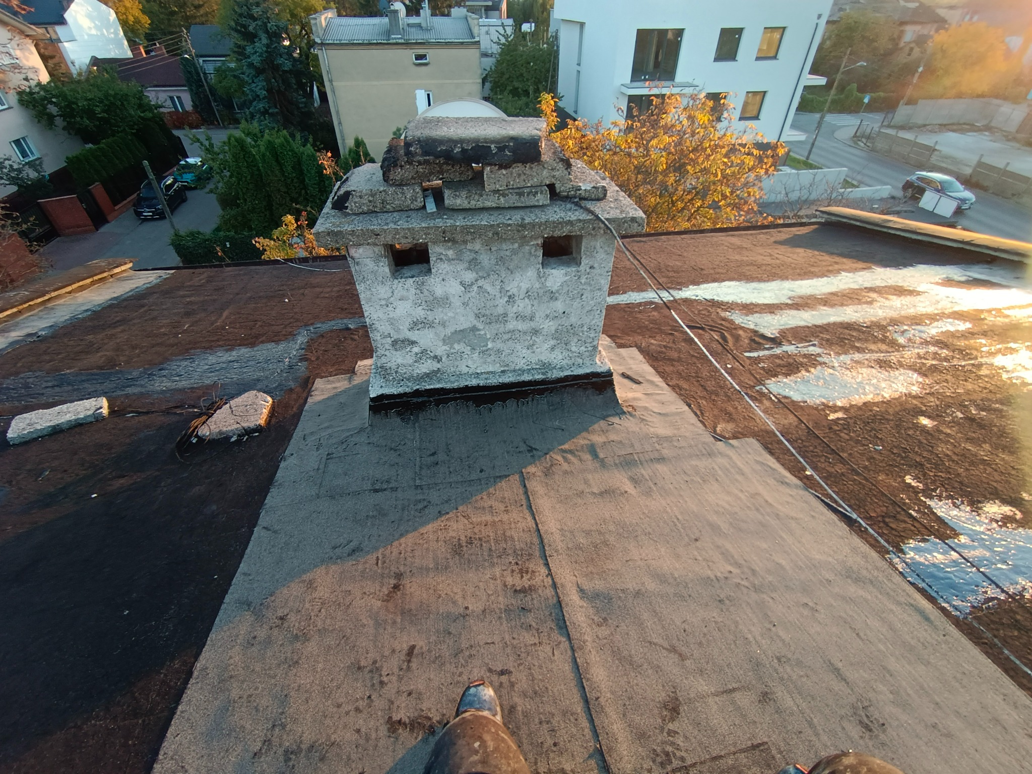 Widok z góry na płaski dach pokryty papą, z betonowym kominem z uszkodzeniami, częściowo naprawiony, w tle zabudowa mieszkalna i drzewa w jesiennym słońcu.