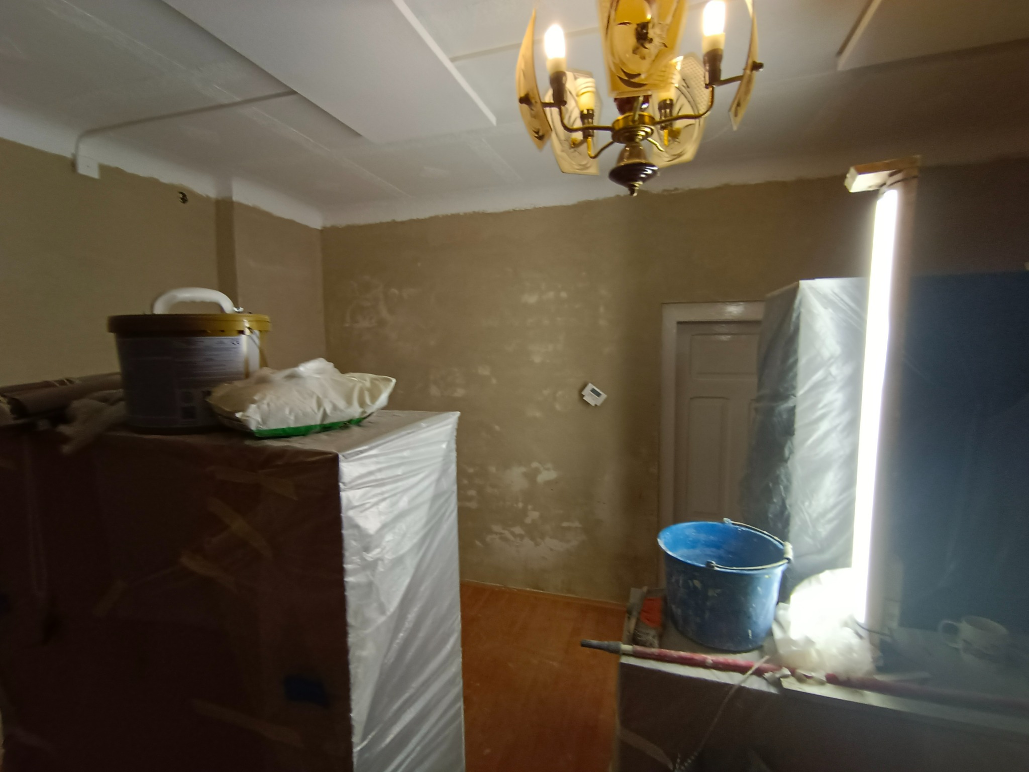 Pokój w trakcie tynkowania ścian, zabezpieczone meble folią, widoczne wiadro, pędzel i lampa halogenowa.