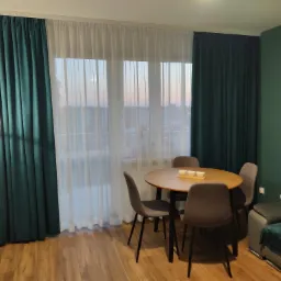 Wnętrze pokoju z ciemnozielonymi, grubymi zasłonami i białymi, prześwitującymi firankami na oknach, okrągły stół z czterema krzesłami i zastawą, widoczna lampa podłogowa i fragment sofy.