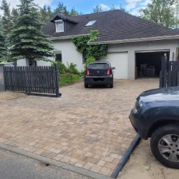 Nowo ułożona kostka brukowa w odcieniach beżu i brązu prowadząca do domu z szarym ogrodzeniem i bramą, z zaparkowanym czarnym samochodem Skoda Citigo i częściowo widocznym ciemnoniebieskim...