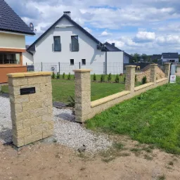 Nowoczesne ogrodzenie z piaskowca z wbudowaną skrzynką na listy, widok z zewnątrz na trawnik, dom i ogrodzenie panelowe w tle, w słoneczny dzień.