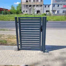 Nowoczesna, szara furtka z poziomymi panelami, zamontowana przy ogrodzeniu z siatki na tle miejskiego krajobrazu z budynkami i drogą.