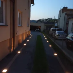 Oświetlona droga dojazdowa do domu z wbudowanymi lampami gruntowymi emitującymi ciepłe światło, widoczne budynki w tle.