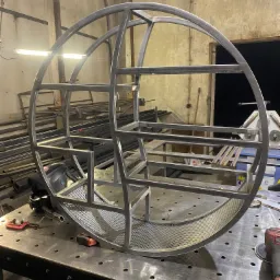 Metalowa konstrukcja w kształcie kuli z wewnętrznymi półkami, wykonana ze stalowych profili, widoczna na stole spawalniczym w warsztacie.