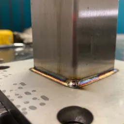 Spawanie metodą TIG: zbliżenie na spoinę łączącą kwadratowy profil ze stalową płytą, widoczne charakterystyczne kolory przegrzanego metalu.