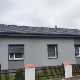 Dom jednorodzinny z nowym, ciemnoszarym dachem z blachodachówki, widoczne okna z roletami zewnętrznymi i numer domu 35, ogrodzenie drewniane z elementami ceglanymi.