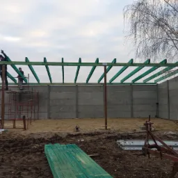 Budowa konstrukcji z drewnianych belek na betonowych fundamentach, widoczni pracownicy na rusztowaniu, ziemisty teren budowy.