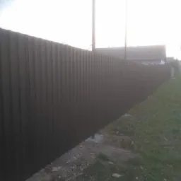 Długi, metalowy płot z blachy falistej w kolorze grafitowym, oddzielający posesję od drogi, widoczny z perspektywy bocznej, z fragmentem trawnika i słupem energetycznym w tle.