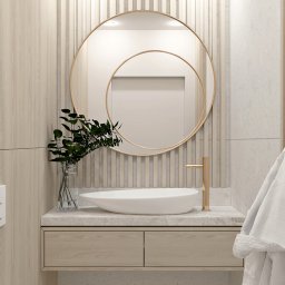 ATKA Architektura Wnętrz Agata Tokarz - Jasna łazienka z okrągłym lustrem z podwójną ramą, białą umywalką nablatową, złotą baterią i drewnianymi elementami wykończeniowymi; szklany wazon z zielonymi liśćmi dodaje świeżości.