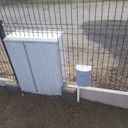 Paweł Miśkiewicz - Szara skrzynka sterownicza i czujnik automatyki bramy Nice zamontowane przy ogrodzeniu z siatki, widoczne betonowe elementy montażowe.