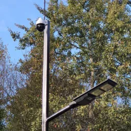 Słup oświetleniowy z kamerą obrotową, zamontowany na tle drzew z zielonymi liśćmi i błękitnym niebem.