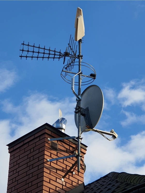 Anteny różnego typu (telewizyjna, satelitarna, radiowa) zamontowane na metalowym maszcie przymocowanym do komina z cegły, na tle błękitnego nieba z chmurami.