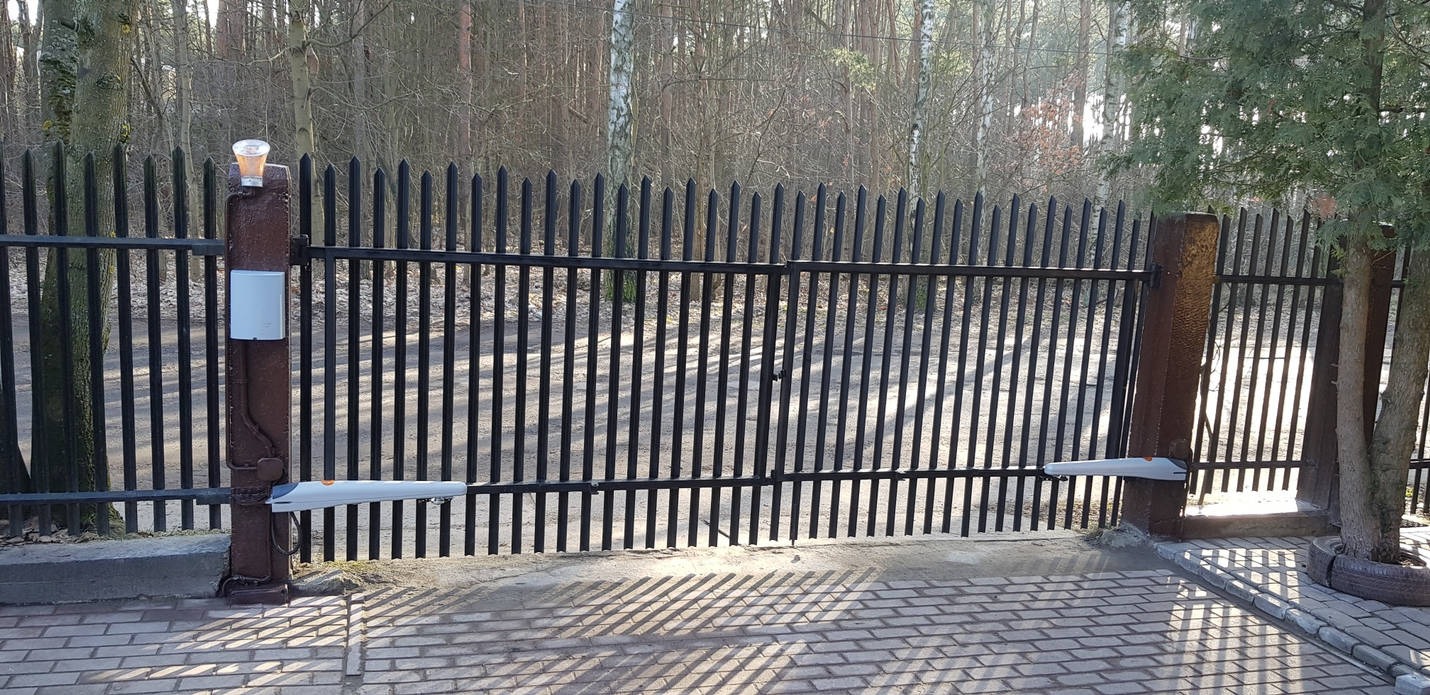 Automatyczna brama wjazdowa z czarnymi prętami i brązowymi słupkami, z widocznymi siłownikami i elementami sterującymi, na tle drzew bez liści i kostki brukowej.
