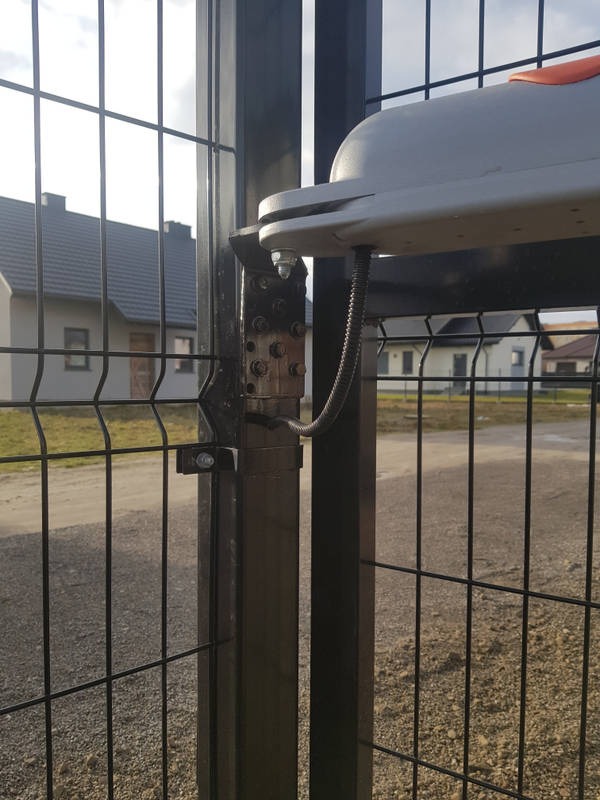 Mechanizm napędu bramy zamontowany na metalowym słupku ogrodzenia panelowego, widoczne śruby mocujące i przewód zasilający, w tle zabudowa jednorodzinna.