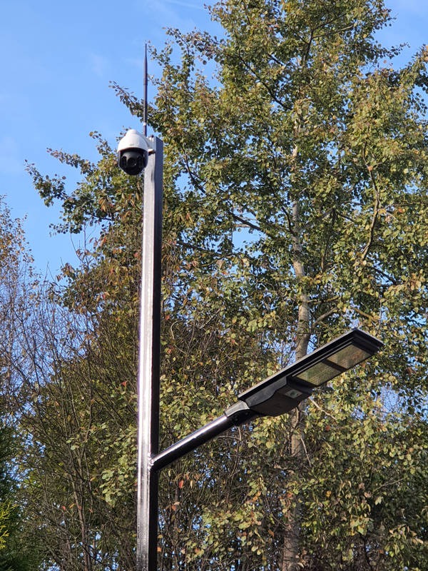 Słup oświetleniowy z kamerą obrotową, zamontowany na tle drzew z zielonymi liśćmi i błękitnym niebem.