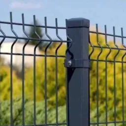 Fragment grafitowego ogrodzenia panelowego z kwadratowym słupkiem mocującym, na tle zieleni i błękitnego nieba.