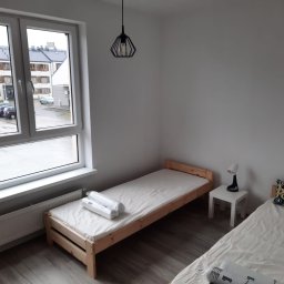 RentPlanet Professionals sp. z o.o. - Pokój z dwoma pojedynczymi łóżkami z drewnianymi ramami, białymi materacami i pościelą, widok z okna na osiedle mieszkaniowe w pochmurny dzień, nowoczesna lampa wisząca.