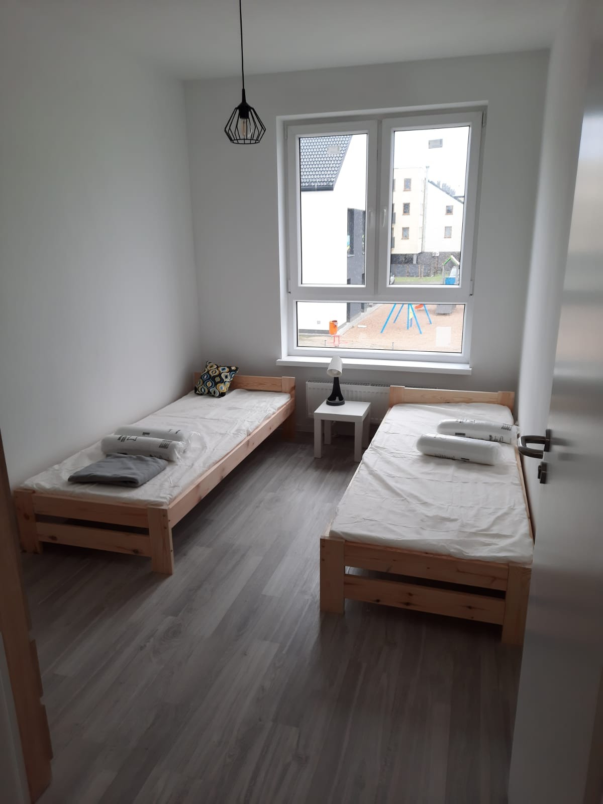 Pokój z dwoma pojedynczymi łóżkami z drewnianymi ramami, białymi pościelami i ręcznikami, widok z okna na plac zabaw, lampa na stoliku nocnym i geometryczna lampa wisząca.