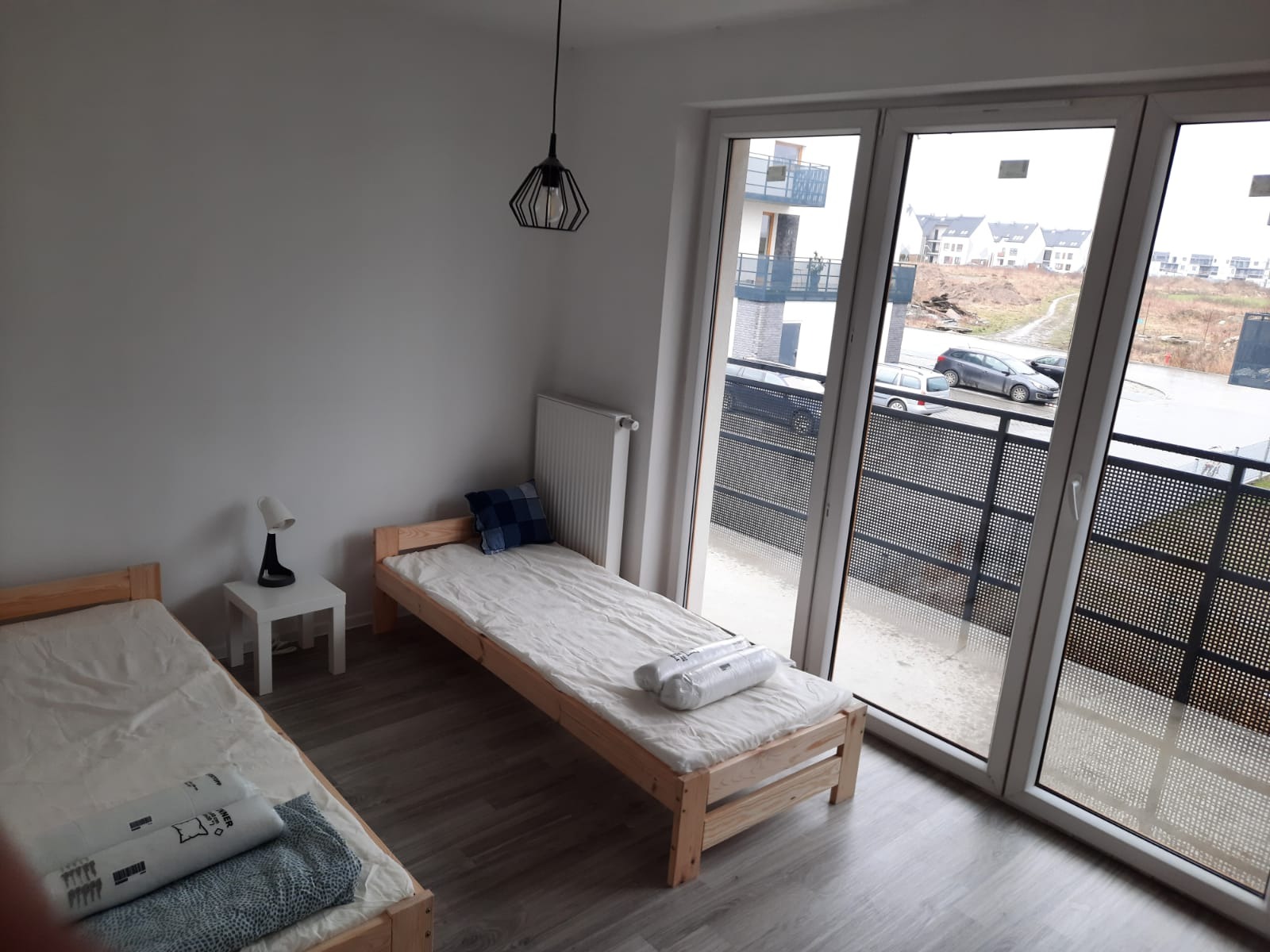 Pokój z dwoma pojedynczymi łóżkami, widok z okna na osiedle mieszkaniowe, minimalistyczna lampa wisząca i lampka nocna na białym stoliku.