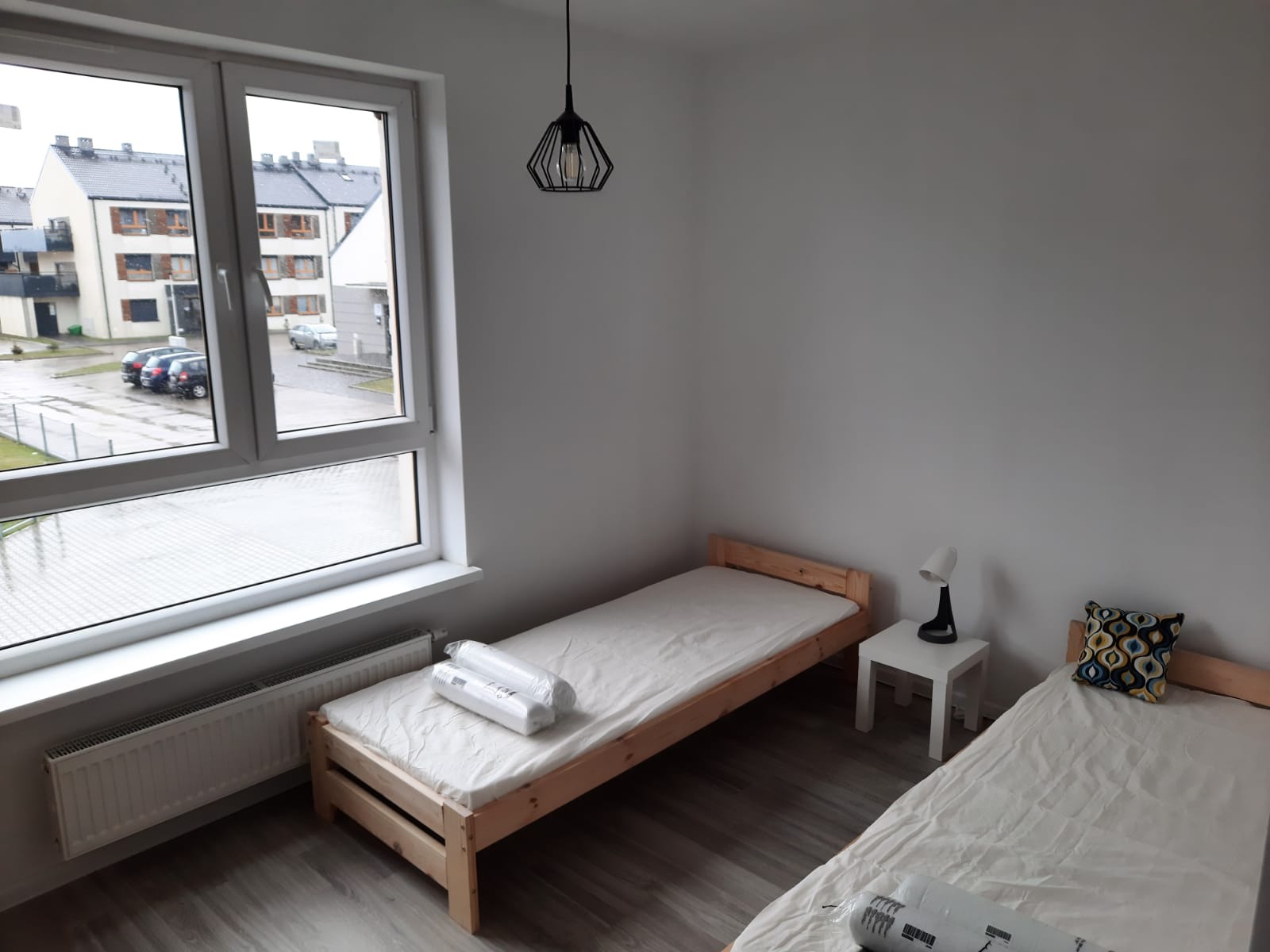 Pokój z dwoma pojedynczymi łóżkami z drewnianymi ramami, białymi materacami i pościelą, widok z okna na osiedle mieszkaniowe w pochmurny dzień, nowoczesna lampa wisząca.
