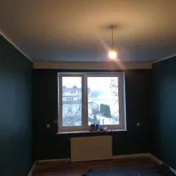 Pokój po świeżym malowaniu ścian na ciemnozielony kolor; widoczne okno z białymi ramami, biały grzejnik i fragment podłogi z panelami; sufit biały z wiszącą żarówką.