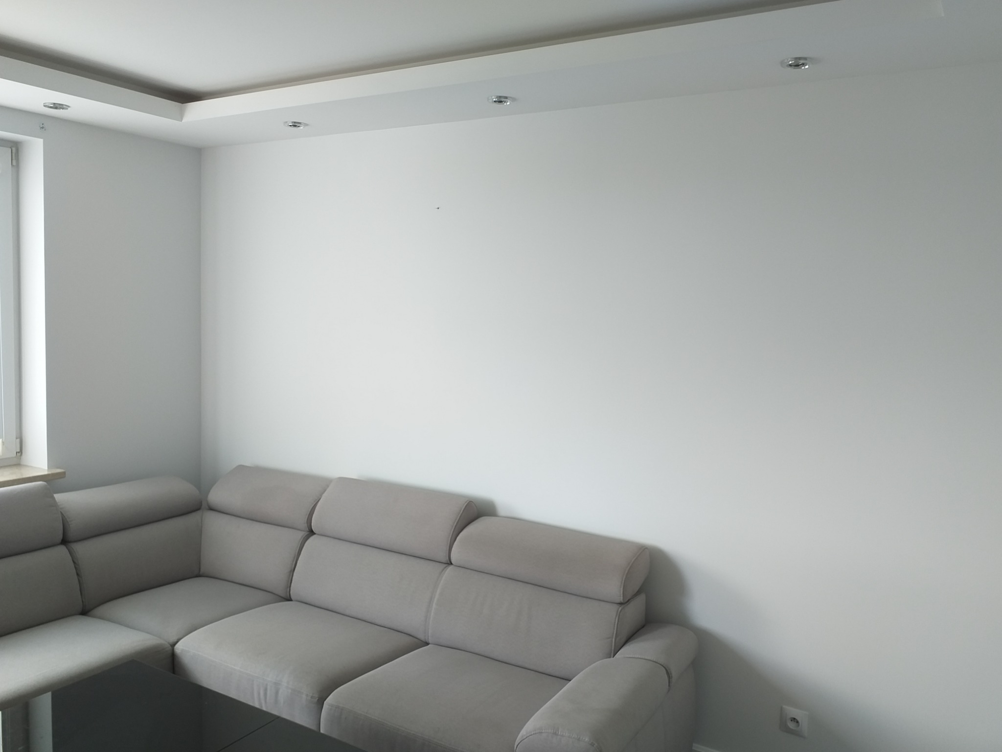 Jasnoszara sofa narożna pod ścianą świeżo pomalowaną na biało, widoczne gniazdko elektryczne i okno w tle.