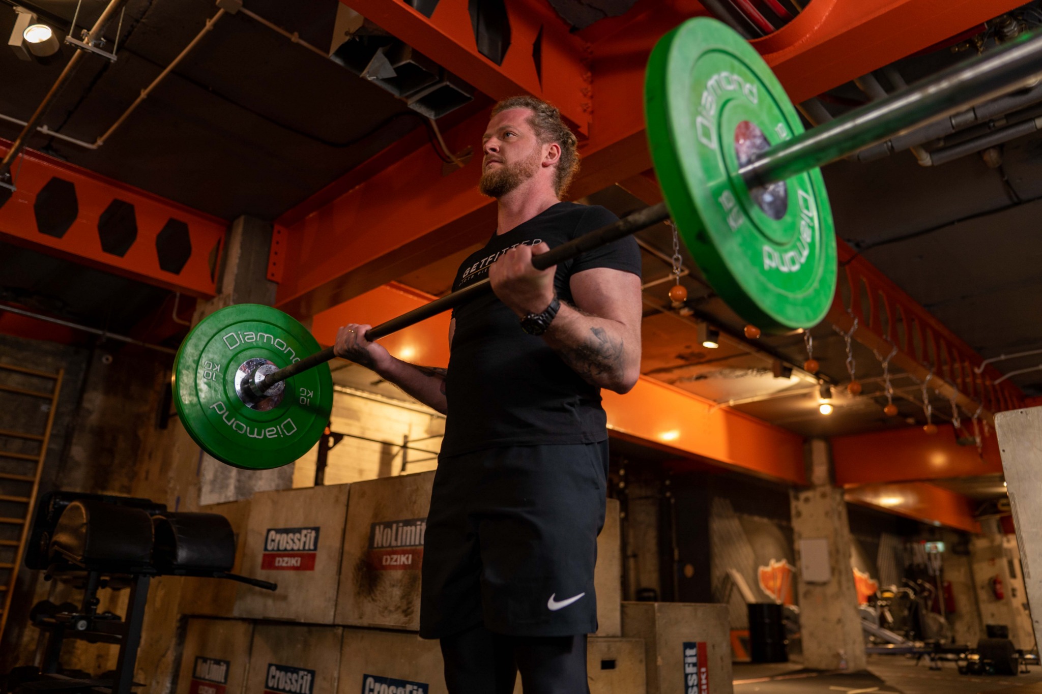 Mężczyzna z brodą podnosi sztangę z zielonymi obciążeniami w siłowni CrossFit z betonowymi ścianami i drewnianymi skrzyniami w tle.