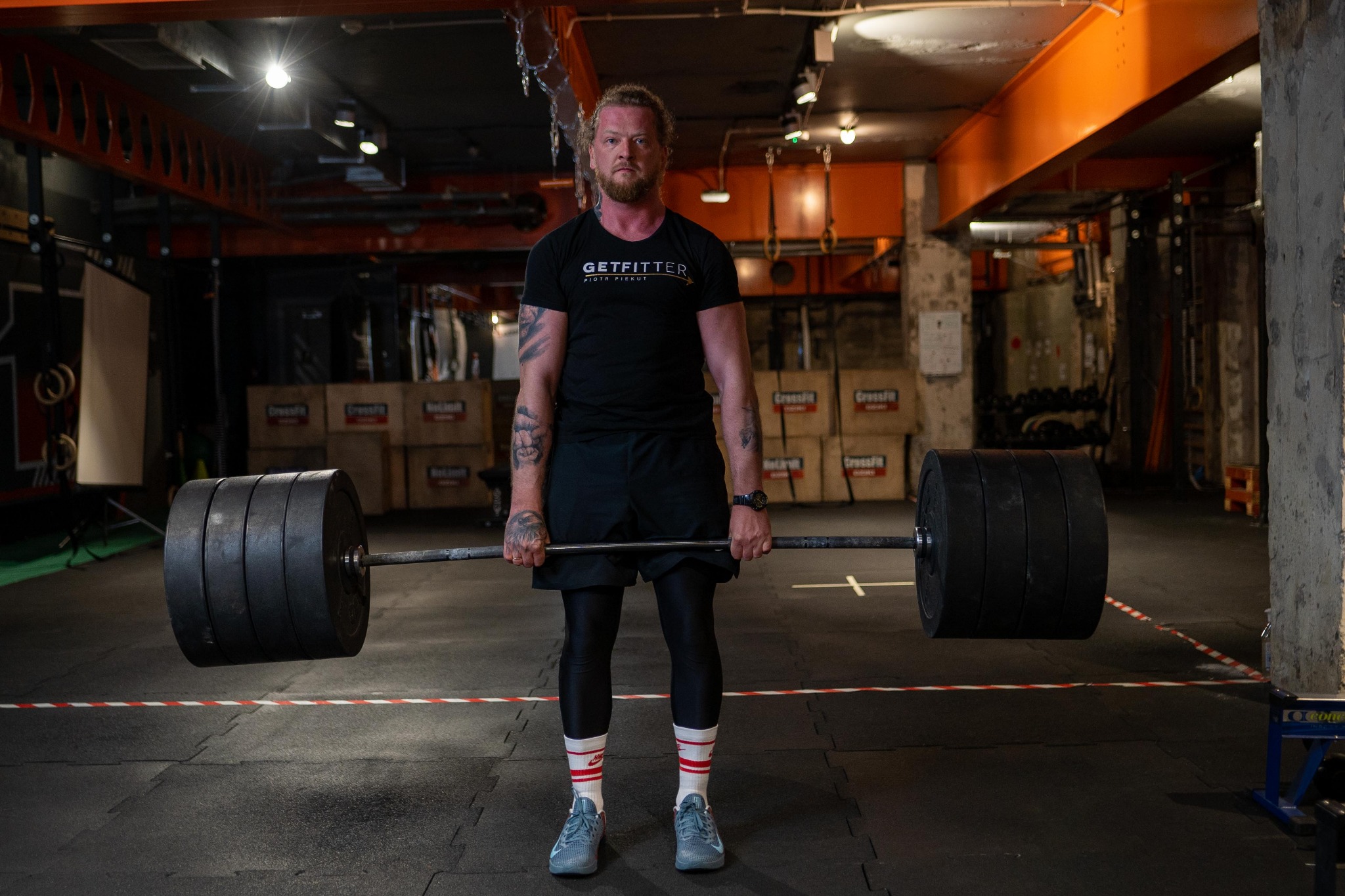 Mężczyzna z brodą i tatuażami, w czarnej koszulce z logo, przygotowuje się do podniesienia sztangi z dużym obciążeniem na siłowni Crossfit