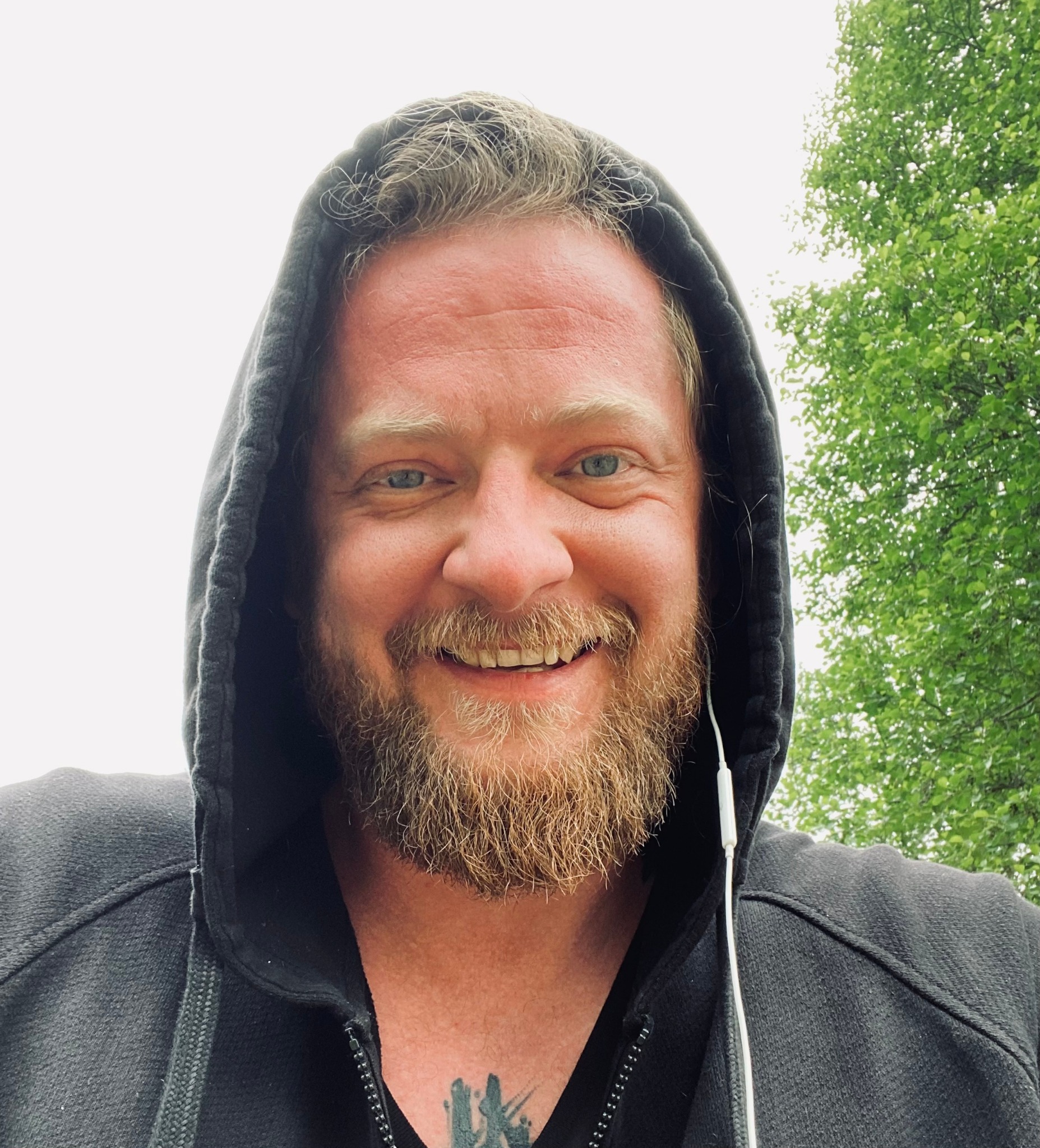 Portret uśmiechniętego mężczyzny w bluzie z kapturem, z brodą i słuchawkami, na tle zieleni w Warszawie.