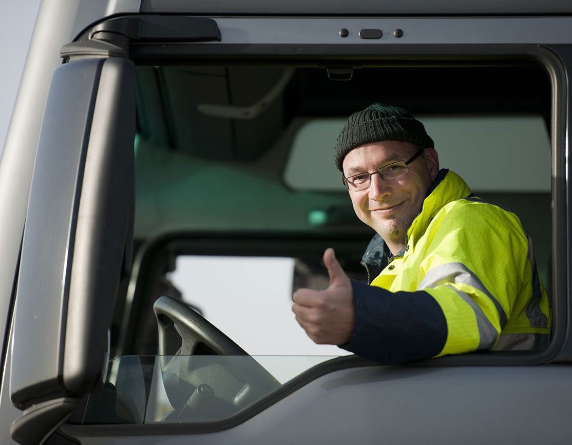 Uśmiechnięty kierowca w czapce i okularach, ubrany w żółtą kurtkę odblaskową, wystawia kciuk z kabiny szarej ciężarówki.