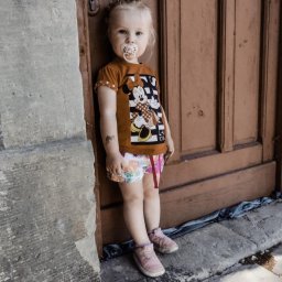 Katarzyna Rudowiecka - Dziewczynka z blond włosami i smoczkiem w ustach, oparta o drewniane drzwi i kamienną ścianę; ubrana w pomarańczową koszulkę z Minie i wzorzyste spodenki.