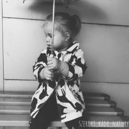 Katarzyna Rudowiecka - Dziewczynka w czarno-białym płaszczu w zwierzęcy wzór, siedząca na schodach i trzymająca parasol, spogląda w lewo, zdjęcie w odcieniach szarości.